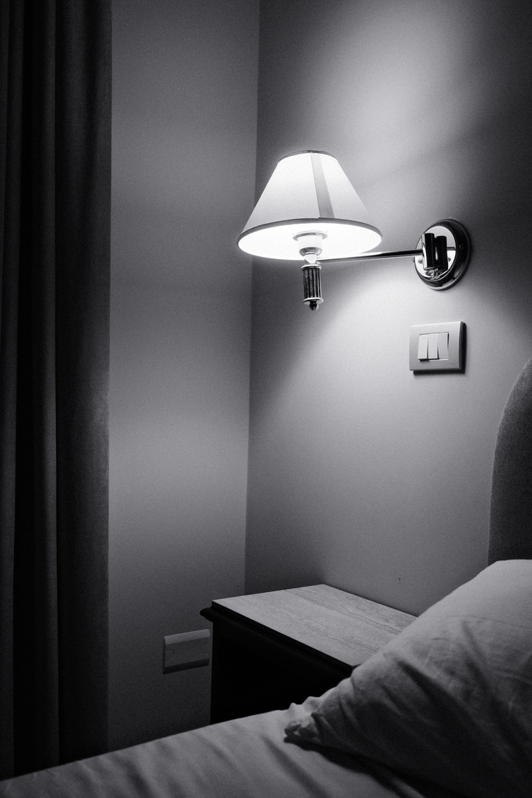Вместо прикроватной тумбы у стены закреплены узкая полка глубиной двадцать сантиметров и настенный светильник на кронштейне